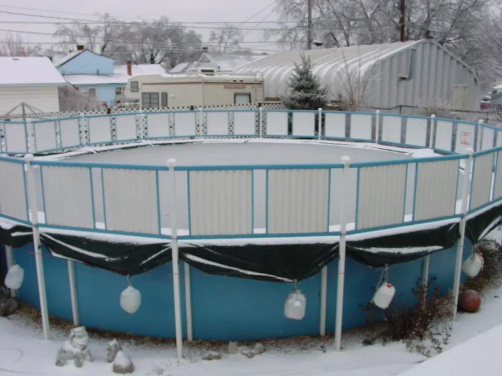 Photo of an above-ground pool closed for winter under snow, its cover weighted with hanging water jugs