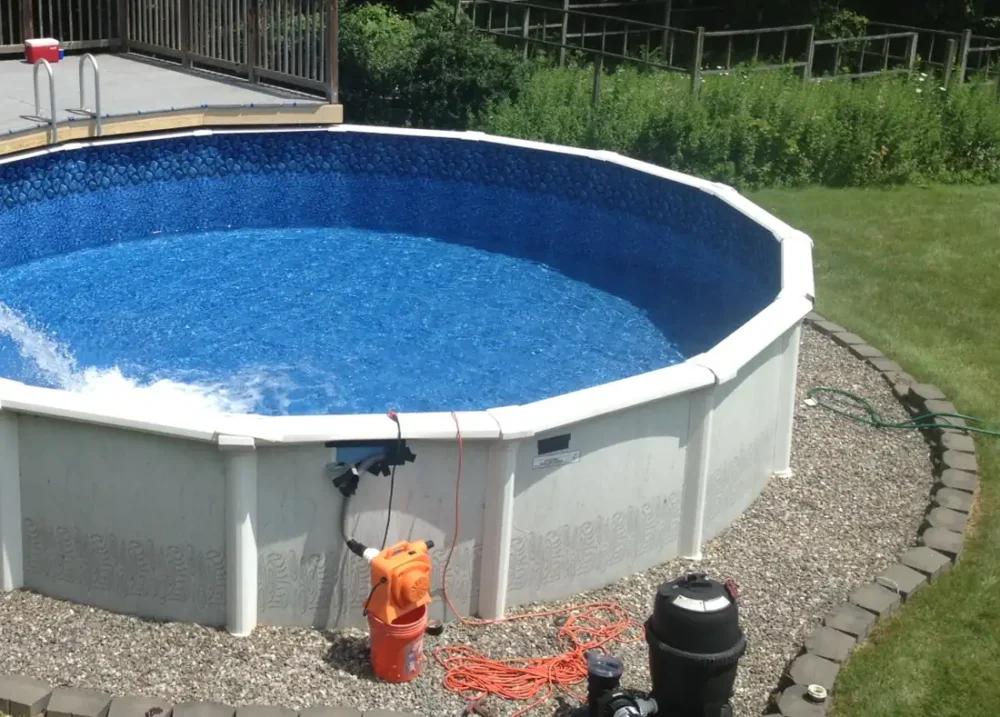 Photo of an above-ground pool being filled with water, with its sand filter and pump on the gravel