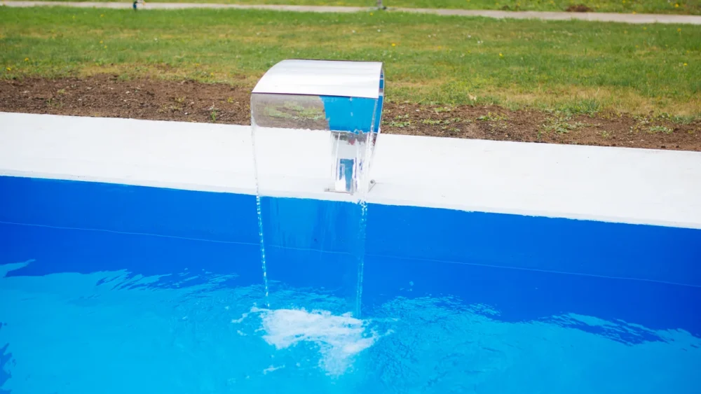 Photo of a backyard pool with clear blue water and a stainless steel water feature pouring in from the coping