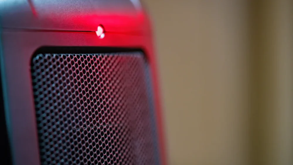Close-up photo of a ceramic electric space heater grille and its glowing red power light against a beige wall