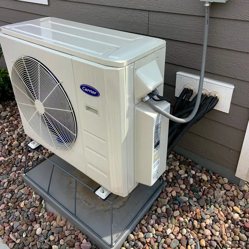 Photo of a ductless mini-split heat pump outdoor unit on a stand beside a house, with refrigerant lines