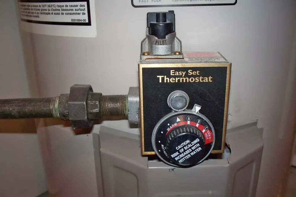 Close-up photo of a gas water heater thermostat dial with a scald caution label and the pilot control above it