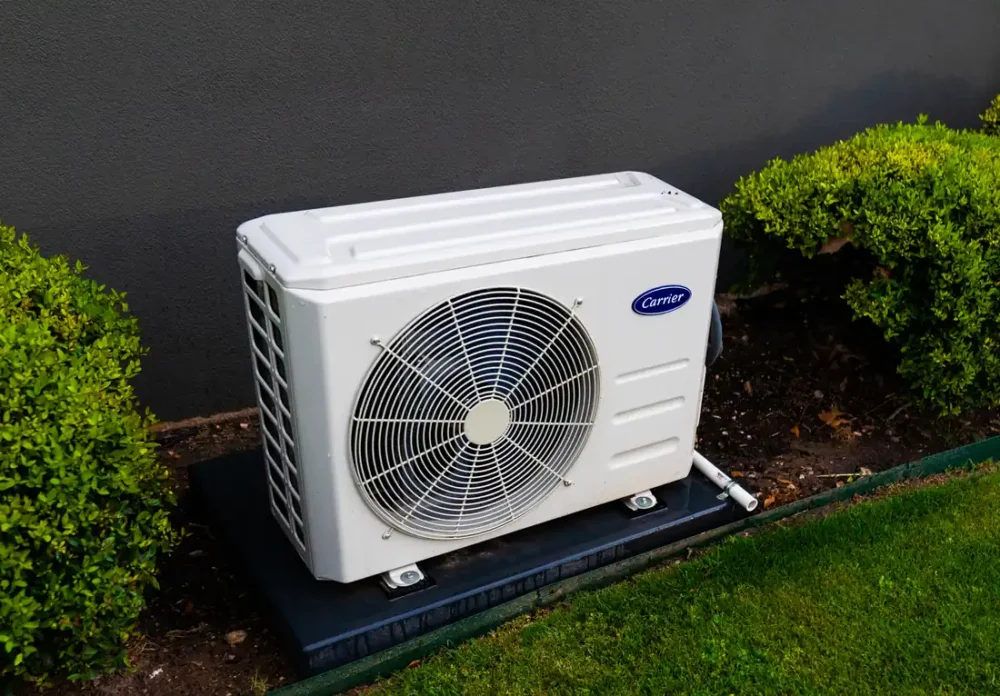 Photo of a white heat pump outdoor unit on a dark pad beside a gray house wall between green shrubs