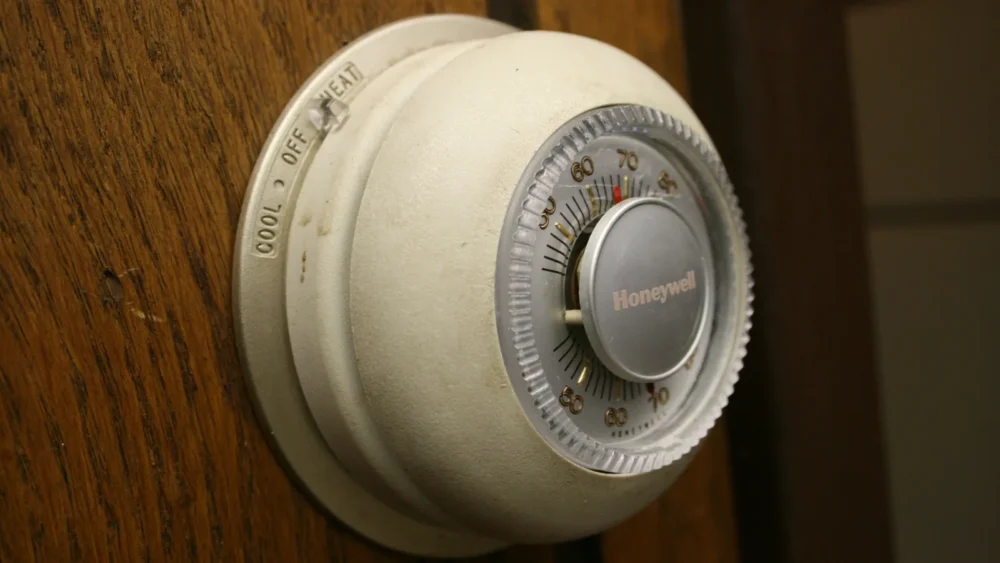 Photo of a classic round Honeywell dial thermostat with a heat, off and cool switch on a wood-paneled wall