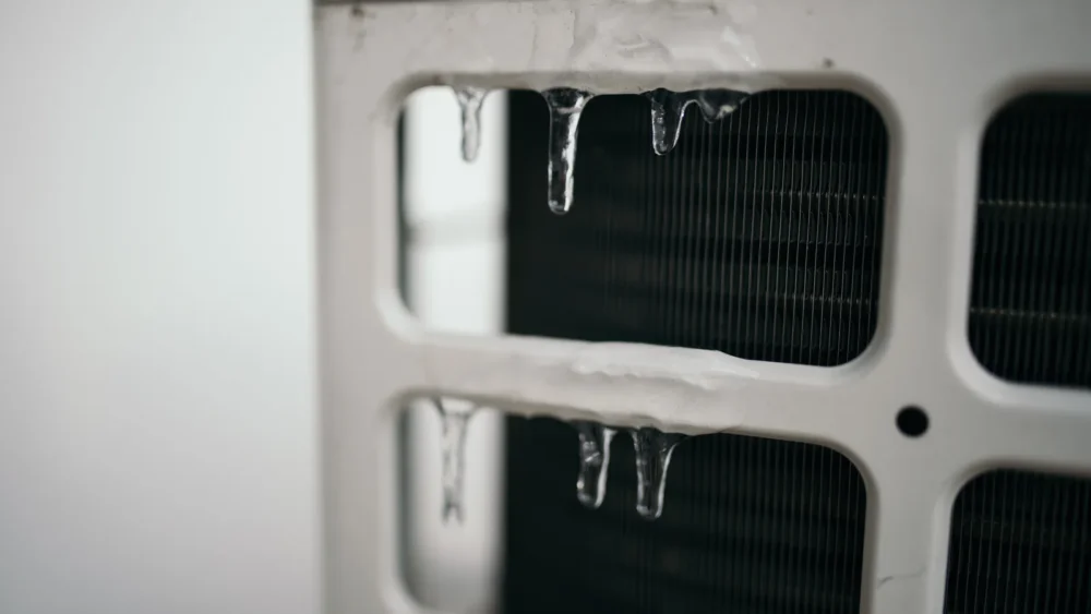 Close-up photo of icicles hanging from the grille of a heat pump outdoor unit, with the coil fins behind