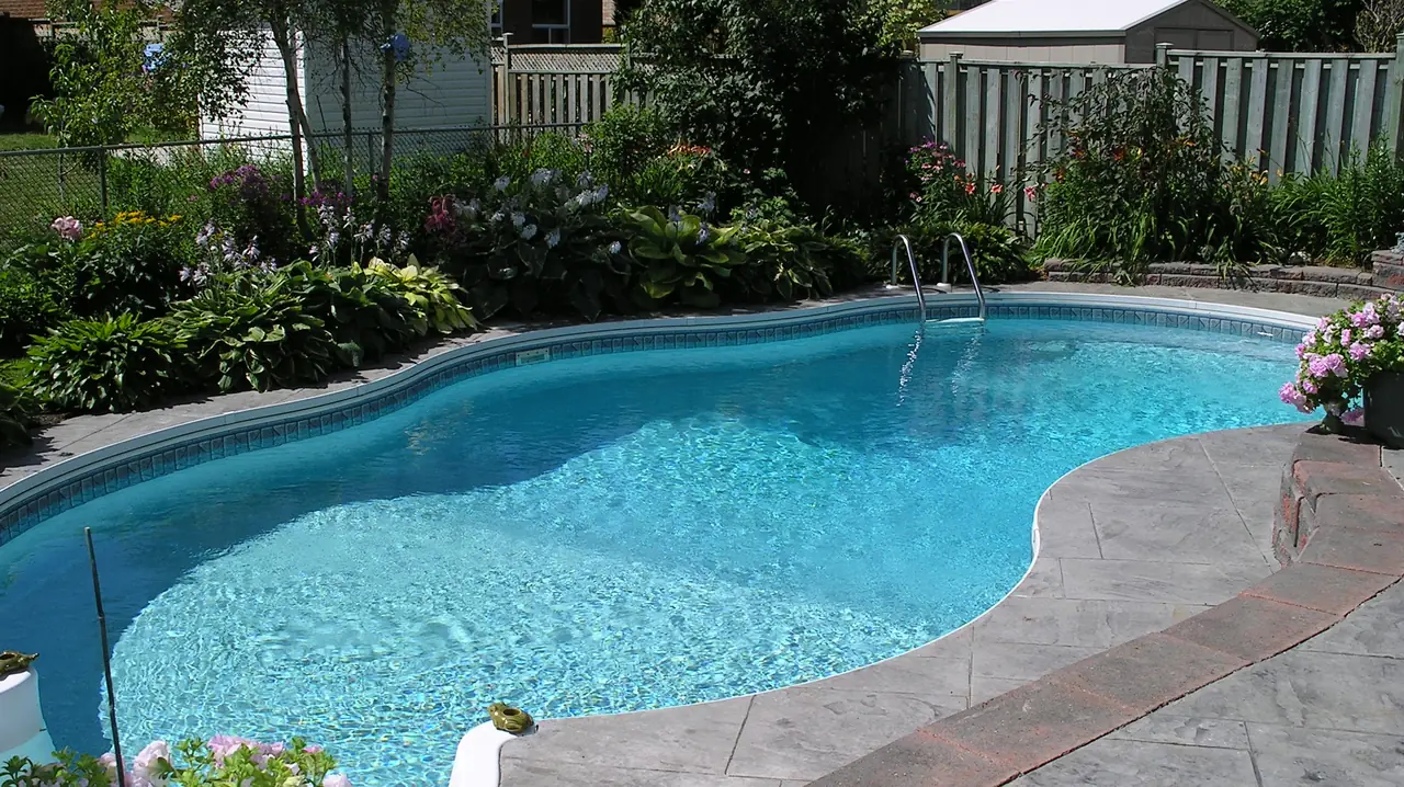 Photo of an open backyard in-ground pool with clear blue water, a ladder and flower beds along a wooden fence